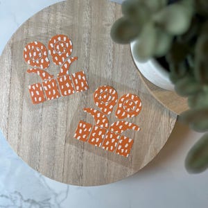 May include: Two orange and white patterned vinyl decals with a swirl design. The decals are on a wooden surface.