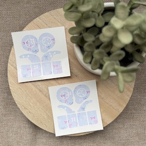 May include: Two sheets of light blue nail art stickers with a bow and floral design. The stickers feature a variety of shapes, including bows and floral patterns in pink and white. The stickers are on a wooden tray with a small potted plant.
