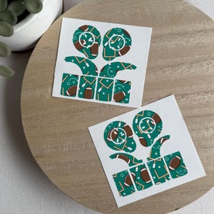 May include: Two white square stickers with a football theme. Each sticker features a football helmet design, footballs, and play diagrams in green, brown, and white. The stickers are on a wooden surface.