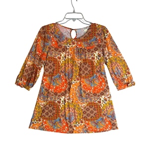 May include: A long-sleeve tunic top featuring a vibrant orange, yellow, and blue paisley pattern. The garment has a round neckline with a small keyhole cutout at the center, three-quarter length sleeves, and a relaxed, straight-cut silhouette.