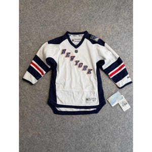 May include: A white Reebok New York Rangers hockey jersey with navy blue and red accents. The front features the text NEW YORK in blue and red lettering, a V-neck collar, and long sleeves with red and navy stripes. A retail tag is attached to the side.