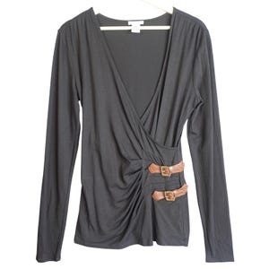以下が含まれることがあります： 深いVネックとラップスタイルが特徴の黒い長袖トップス。衣服の側面には、それぞれゴールドトーンの金属製バックルで固定された2本の茶色のフェイクレザー製ストラップがあしらわれており、生地に繊細なギャザーのディテールが施されています。