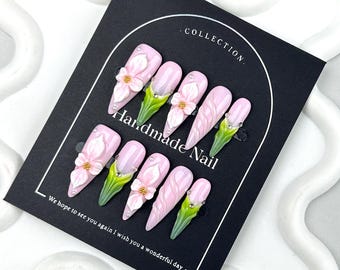 Könnte beinhalten: Ein Set aus zehn künstlichen Nägeln mit hellrosa Basis, verziert mit 3D-weißen Blumen, grünen Akzenten und kleinen Strasssteinen. Die Nägel werden auf einer schwarzen Karte mit dem Text "Handmade Nail" und "Collection" präsentiert.