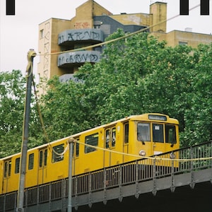 May include: A yellow subway train travels on an elevated track. The train is labelled "11" and "Invalidenstraße". The background includes a building with graffiti and lush green trees. The word "Himmel" is at the top.