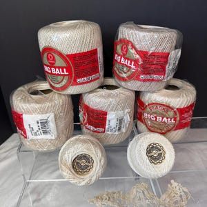 May include: A collection of beige crochet thread balls in various sizes. The larger balls are labeled "BIG BALL" with red and white labels. Smaller balls are also visible, with a "20" label. The thread is tightly wound and ready for crafting.