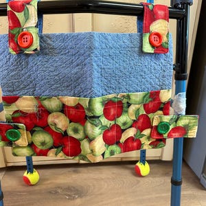 May include: A blue fabric walker bag with a green and red apple print. The bag has two straps with green buttons and a blue fabric pocket on the front.