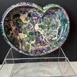 May include: Heart-shaped decorative bowl with a marbled design in shades of green, purple, and white. The bowl is displayed on a clear acrylic stand, showcasing its unique shape and color variations. The bowl's interior features a swirling pattern.