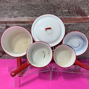 May include: A collection of vintage enamelware pots and a lid. The pots are white with red rims and handles. The lid is white with a red handle. The pots are arranged on a wire rack.