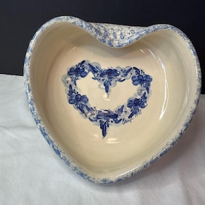 May include: Heart-shaped ceramic bowl with a cream-colored interior and a blue floral design in the center. The rim and outer edge feature a blue speckled pattern. The bowl has a wavy edge and a signature in the bottom right corner.