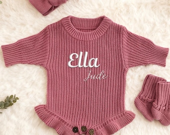 May include: A dusty rose knitted baby outfit set, including a romper with the name "Ella Jude", a matching headband, and booties. The romper has a ruffled bottom and button details. A teddy bear is in the background.
