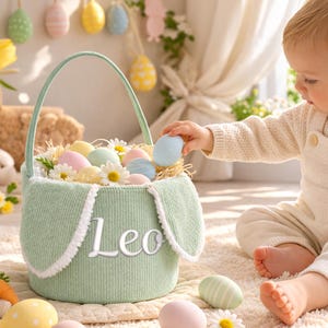May include: A light green Easter basket with bunny ear accents, filled with pastel-colored eggs and daisies. The basket has the name "Leo" embroidered on the front. A baby in cream-colored overalls reaches for an egg. Easter decorations are in the background.