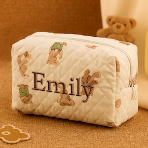 May include: Cream quilted cosmetic bag with a zip closure. The bag is embroidered with the name "Emily" in brown thread and features repeating teddy bear illustrations. A small, bear-shaped patch is visible.