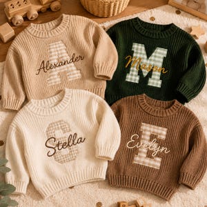 May include: Four long-sleeved knit jumpers in beige, dark green, cream, and brown. Each jumper features a large, centred gingham-patterned letter patch with a name embroidered in cursive script over the letter: Alexander, Mason, Stella, and Evelyn.