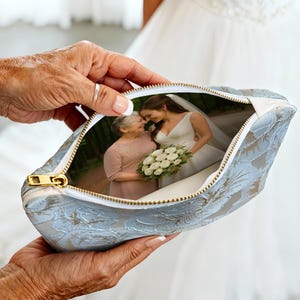 May include: A light blue floral patterned pouch with a gold zipper, holding a photograph of a bride and another woman. The pouch is held by a person with aged hands, and the background shows a white wedding dress.