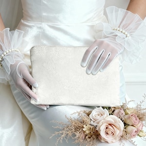 May include: A white clutch with a floral pattern is held by a person wearing sheer white gloves and ruffled wrist cuffs adorned with pearls. A bouquet of roses and dried flowers rests below.