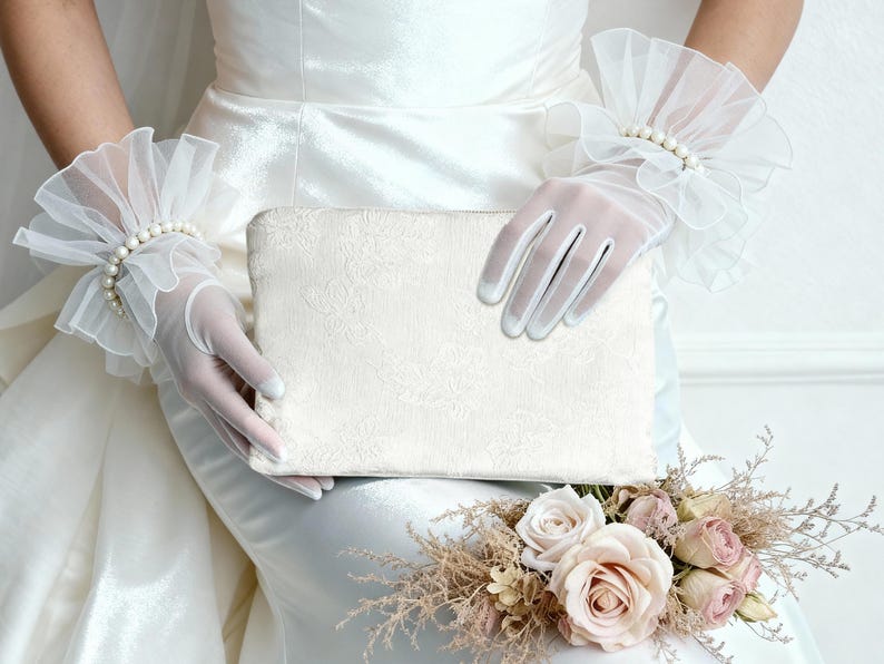 May include: A white clutch with a floral pattern is held by a person wearing sheer white gloves and ruffled wrist cuffs adorned with pearls. A bouquet of roses and dried flowers rests below.