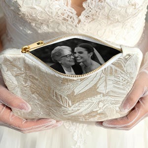 May include: Cream-colored clutch with a gold zipper, featuring a floral pattern. The open zipper reveals a black and white photograph of two smiling women. The clutch is held by a person wearing a white dress and lace gloves.