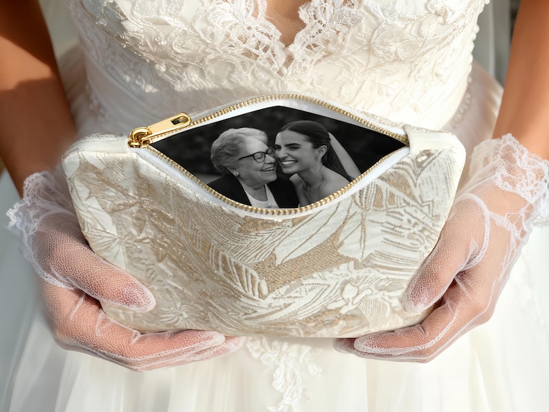 May include: Cream-colored clutch with a gold zipper, featuring a floral pattern. The open zipper reveals a black and white photograph of two smiling women. The clutch is held by a person wearing a white dress and lace gloves.
