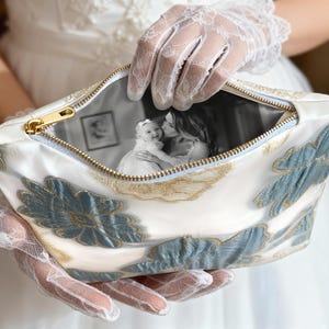 May include: A white cosmetic bag with a gold zipper and blue and gold floral embroidery. The bag's interior displays a black and white photograph of a baby and a woman. The bag is held by a person wearing white lace gloves.