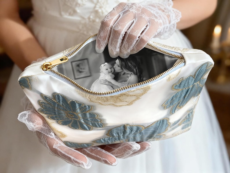 May include: A white cosmetic bag with a gold zipper and blue and gold floral embroidery. The bag's interior displays a black and white photograph of a baby and a woman. The bag is held by a person wearing white lace gloves.