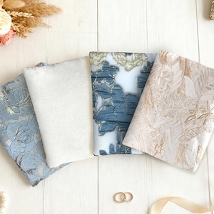 May include: Four folded fabric samples in blue, white, and gold floral patterns are arranged on a white wooden surface. A bouquet of pink and white roses, along with a strand of pearls, and two gold rings are also present.
