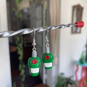 May include: A pair of green and red bottle-shaped earrings with silver hooks. The bottles have a white label and a red cap.