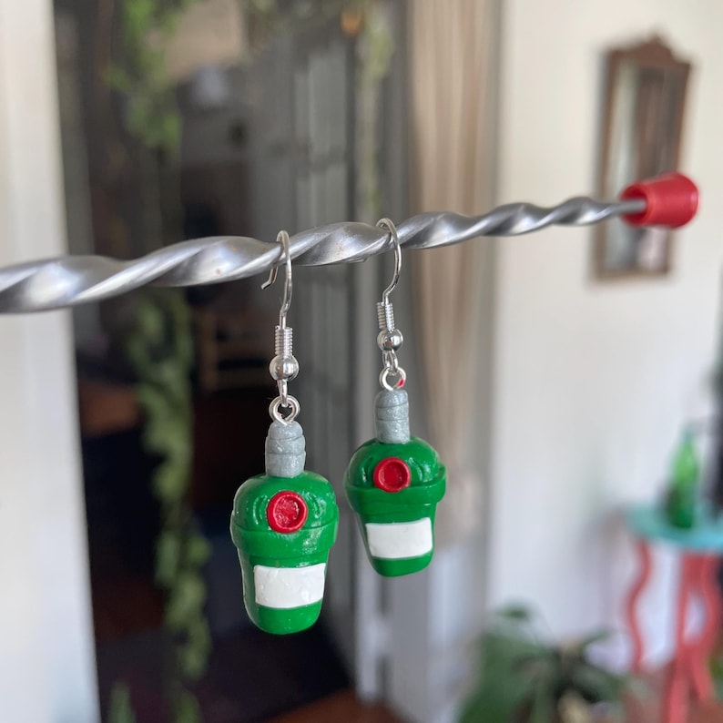 May include: A pair of green and red bottle-shaped earrings with silver hooks. The bottles have a white label and a red cap.
