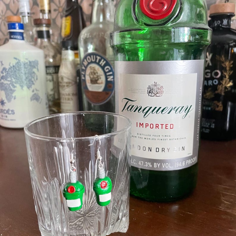 May include: A pair of green glass bottle earrings with silver hooks. The earrings are in a clear glass, resembling a shot glass. The earrings are in front of a bottle of Tanqueray London Dry Gin.