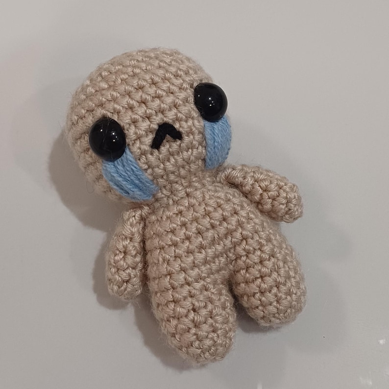 May include: A small crocheted doll with a sad expression. The doll has black eyes, a frown, and blue teardrop-shaped stitches on its cheeks.