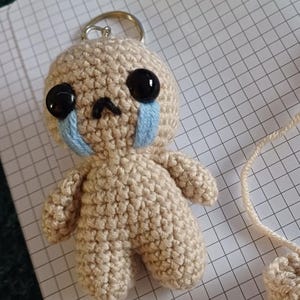 May include: A small crocheted doll keychain with a sad expression. The doll is beige with black eyes and blue tear-shaped accents.