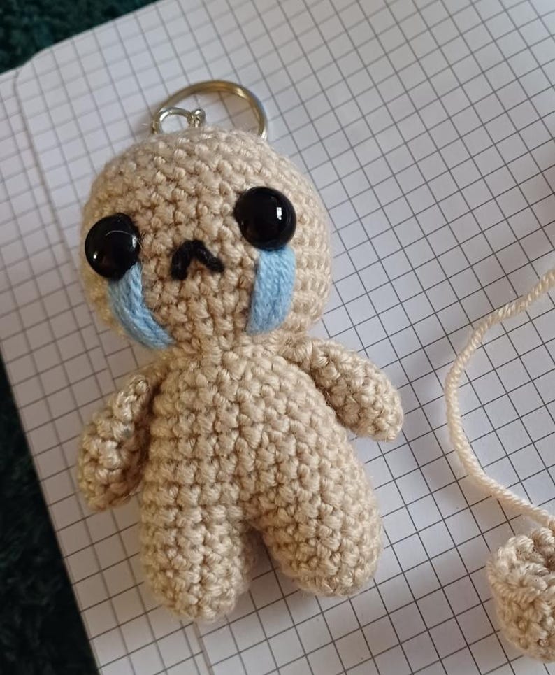 May include: A small crocheted doll keychain with a sad expression. The doll is beige with black eyes and blue tear-shaped accents.