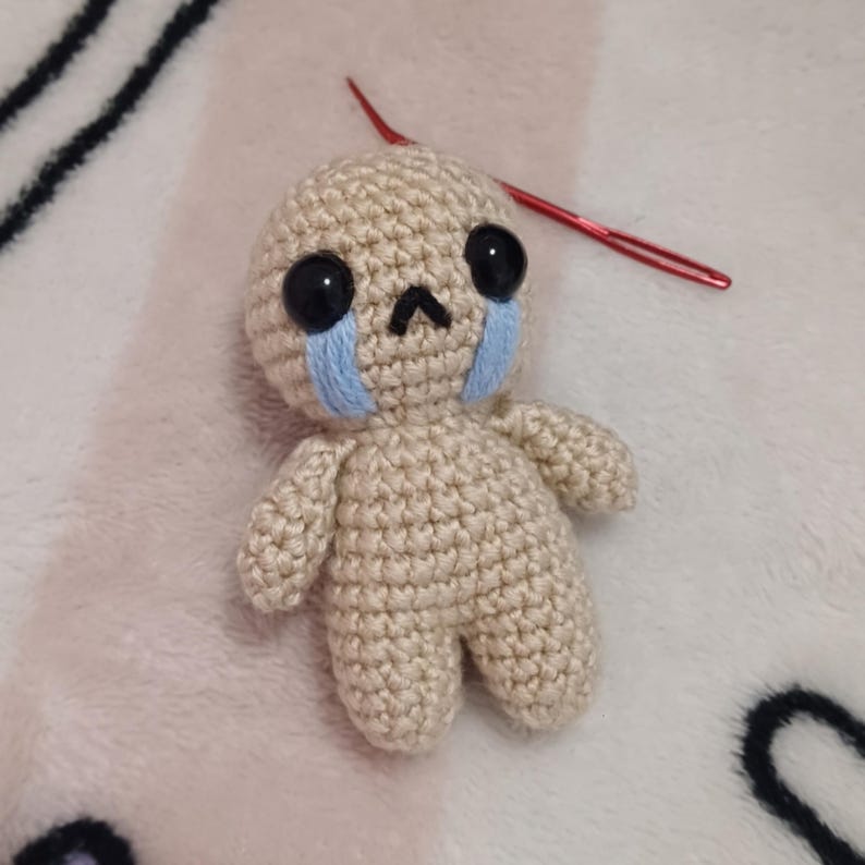 May include: A small crocheted doll with a sad expression. The doll has black eyes, a small nose, and blue embroidered tears. The doll is made of beige yarn and is sitting on a white fuzzy surface.