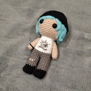 May include: Crocheted doll featuring a black beanie, blue hair, and a white shirt with a graphic design. The doll has grey trousers with patches and black shoes. It rests on a grey, textured surface.