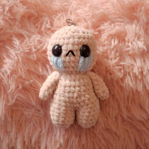 Könnte beinhalten: Eine kleine, beige, gehäkelte Plüschfigur mit rundem Kopf und Körper. Sie hat zwei große, glänzende schwarze Augen, einen kleinen gestickten Schmollmund und hellblaue Garnakzente unter den Augen, die wie Tränen aussehen. Ein Schlüsselring ist befestigt.