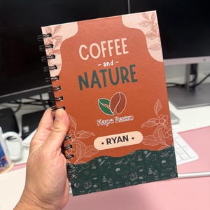 May include: A spiral-bound notebook with a rust-orange cover featuring the text COFFEE - and - NATURE, a coffee bean graphic, the brand name Kape Razzo, and the name RYAN in a white box. The bottom section is dark green with small white icons.