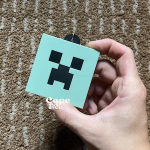 May include: A light blue square keychain with a black Creeper face from the video game Minecraft.