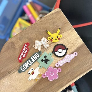May include: A collection of colorful charms featuring various designs, including a Pikachu, a bow, a Minecraft block, a flower, a Pokeball, a chicken, and text that says "Gotta Catch 'Em All!", "Copeland", and "Swiftie".