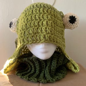 May include: Green crocheted frog hat with a matching scarf. The hat has large white eyes with black pupils and a green crocheted hood.