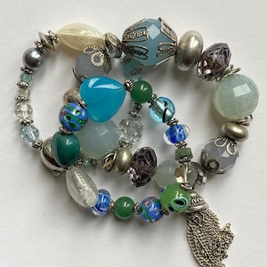 Peut inclure: Plusieurs bracelets de perles avec une variété de perles colorées dans les tons de bleu, vert, argent et transparent. Les bracelets comprennent des perles facettées, rondes et en forme de cœur, avec un pompon argenté sur l'un d'eux. Les perles sont enfilées avec des apprêts en métal argenté.