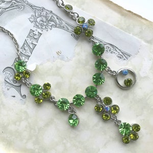May include: A silver-toned necklace with a delicate design. The necklace features clusters of round, green gemstones in varying shades, with a few iridescent blue accents. The gemstones are connected by a fine chain.