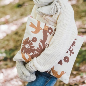 May include: A white canvas tote bag with brown floral and bird illustrations. The bag has a pink zipper and a brown leather strap.