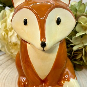 May include: A ceramic fox ornament with a glossy finish. The fox has a reddish-brown body, cream-coloured face and chest, and black eyes. The figurine is sitting upright, with its tail curled to the side. The fox is a decorative item.