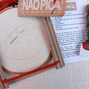 May include: A close-up of a craft kit for embroidery. The kit includes a wooden embroidery hoop with white fabric inside, a needle, red embroidery floss, and a card with instructions in Portuguese. The card reads "NAOPICA PICA" which translates to "Doesn't prick, pricks".