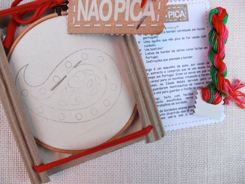 May include: A close-up of a craft kit for embroidery. The kit includes a wooden embroidery hoop with white fabric inside, a needle, red embroidery floss, and a card with instructions in Portuguese. The card reads "NAOPICA PICA" which translates to "Doesn't prick, pricks".