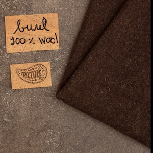 May include: A piece of dark brown felted wool fabric next to two rectangular cork labels. The top label features the handwritten text "burel 100% wool" in black ink, while the bottom label displays a paisley-shaped logo with the word "FOLCLORE".