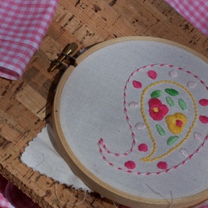 May include: A white embroidery hoop with a paisley design embroidered in pink, yellow, and green thread. The design includes small flowers and leaves.