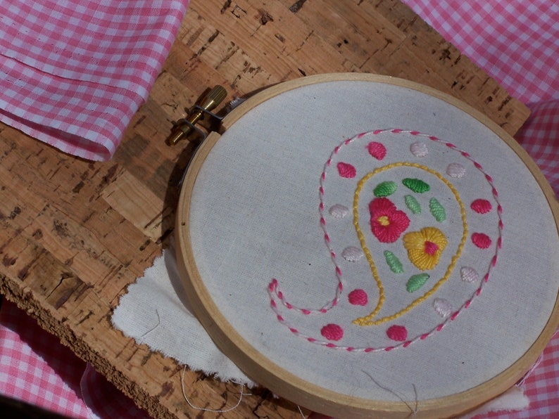 May include: A white embroidery hoop with a paisley design embroidered in pink, yellow, and green thread. The design includes small flowers and leaves.