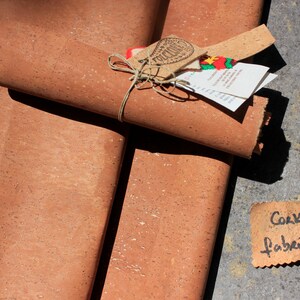 May include: Two rolls of cork fabric, one rolled up and tied with twine, the other unrolled. The fabric is a light brown color with a speckled texture. A small tag with the text "Cork Fabric" is visible in the lower right corner of the image.
