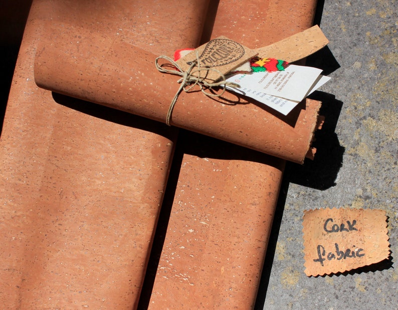 May include: Two rolls of cork fabric, one rolled up and tied with twine, the other unrolled. The fabric is a light brown color with a speckled texture. A small tag with the text "Cork Fabric" is visible in the lower right corner of the image.