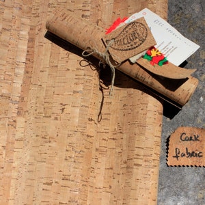 May include: A roll of cork fabric, unrolled and lying on a grey surface. The fabric is a natural brown colour and has a textured surface. A small tag with the text "Cork Fabric" is visible in the lower right corner of the image.